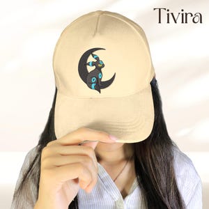 Op de afbeelding: Beige baseballpet met een zwart Umbreon-ontwerp op een halve maan met blauwe details. De pet heeft een gebogen klep en het woord "Tivira" staat rechtsboven gedrukt.
