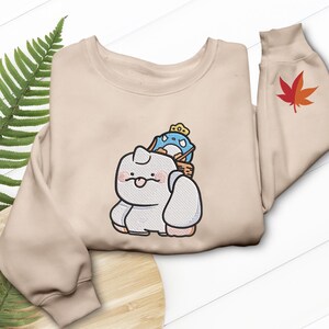 May include: A beige sweatshirt featuring an embroidered cartoon design of a white creature with a blue and yellow character on its back. A red and orange maple leaf is embroidered on the sleeve.