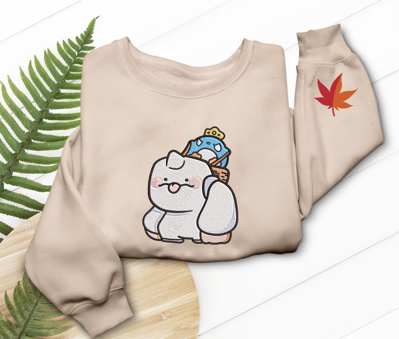 May include: A beige sweatshirt featuring an embroidered cartoon design of a white creature with a blue and yellow character on its back. A red and orange maple leaf is embroidered on the sleeve.