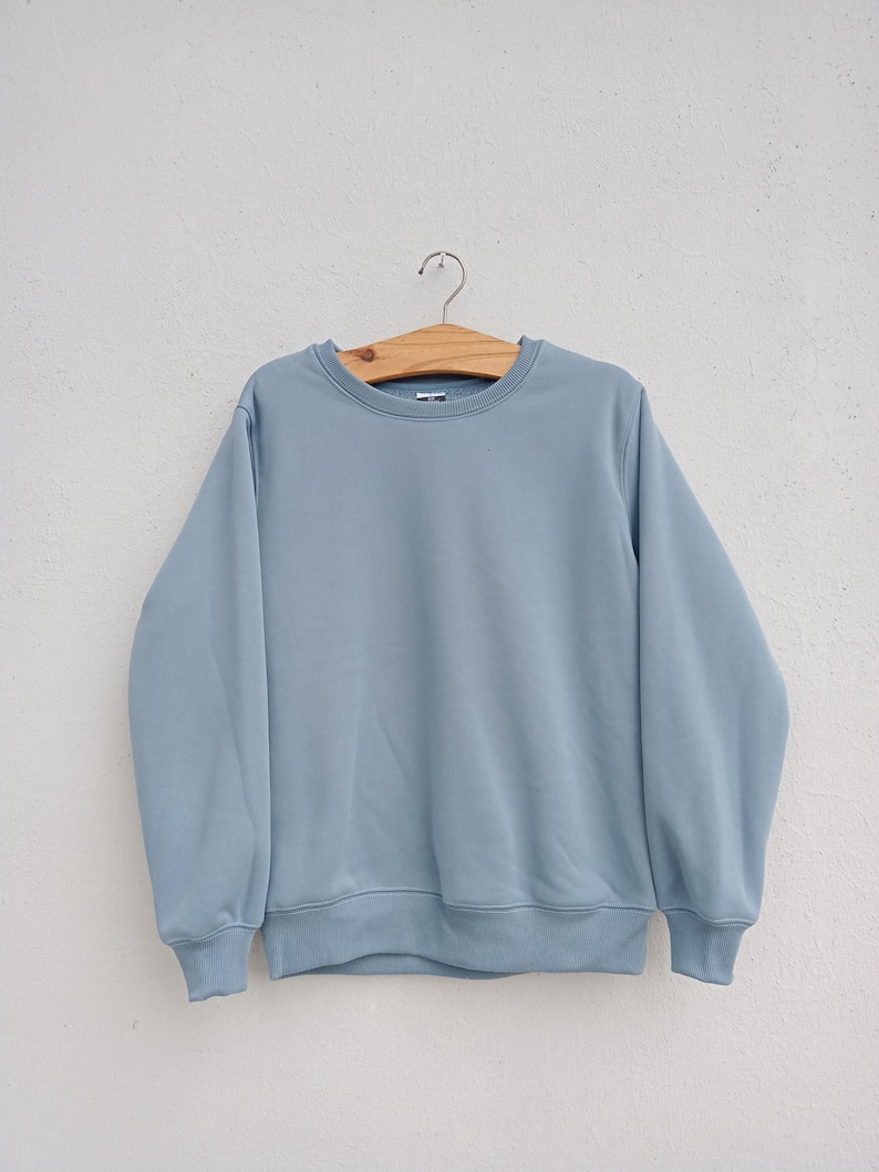 May include: A light blue sweatshirt hangs on a wooden hanger against a white wall. The crew neck sweatshirt has long sleeves and ribbed cuffs and hem. The casual top is a solid color and appears to be made of a soft material.