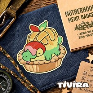 May include: An embroidered patch featuring a green creature with a red heart-shaped head resting inside a golden-brown pie crust, topped with lattice-style pastry strips and a small red leaf, set against a dark blue fabric background.