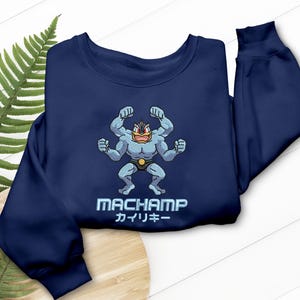 Könnte beinhalten: Marineblaues Sweatshirt mit einem Cartoon-Machamp-Design. Die Machamp-Figur ist hellblau mit einem gelben Gürtel und roten Details. Das Wort "MACHAMP" und japanischer Text sind darunter gedruckt.