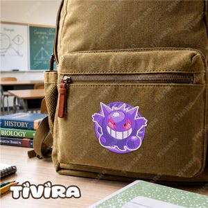 May include: A brown canvas backpack featuring a circular embroidered patch of a purple cartoon creature with red eyes and a wide grin. The backpack has a front zippered pocket and is positioned on a desk with books and pencils nearby.