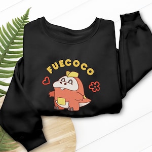 Peut inclure: Sweat-shirt noir avec un personnage de dessin animé brodé et le mot "FUECOCO" en jaune. Le personnage est orange et jaune, avec des graphiques de cœur et de fleur.