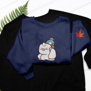 May include: Navy blue sweatshirt featuring an embroidered cartoon cat carrying a blue bird with a crown. A red maple leaf is on the sleeve. The sweatshirt is layered over a black sweatshirt.
