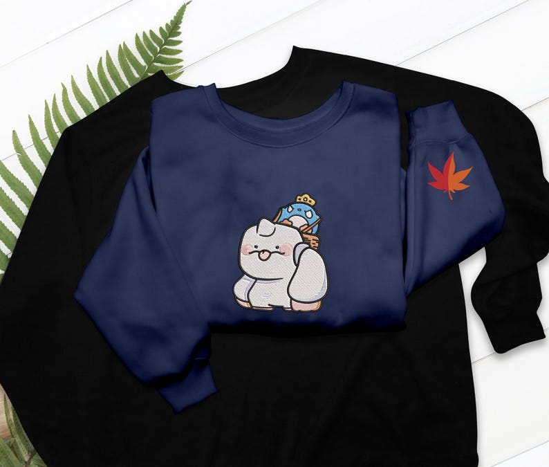 May include: Navy blue sweatshirt featuring an embroidered cartoon cat carrying a blue bird with a crown. A red maple leaf is on the sleeve. The sweatshirt is layered over a black sweatshirt.