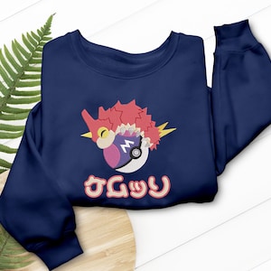 May include: Navy blue sweatshirt featuring an embroidered design of a bird-like creature with red, pink, and white accents, and a stylized Poké Ball. Japanese text is below the design. The sweatshirt has a crew neck and long sleeves.
