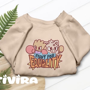 Op de afbeelding: Een beige sweater met ronde hals en een geborduurde afbeelding van twee vechtende stripfiguren met bokshandschoenen, geplaatst boven de tekst "FIGHT FOR EQUALITY" in dikke, gestileerde letters met een gele en rode omlijning.