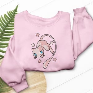 Könnte beinhalten: Ein hellrosa Sweatshirt mit einer Cartoon-Illustration einer rosa katzenartigen Kreatur mit blauen Augen und einem langen Schwanz. Das Design ist von kleinen Blumen umgeben. Das Sweatshirt hat einen Rundhalsausschnitt und lange Ärmel.