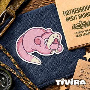May include: A pink creature sticker with a cream-coloured tail tip and open mouth, positioned on a dark blue fabric surface next to a tan card labelled MOTHERHOOD MERIT BADGE and a compass with a green and brown braided cord strap.