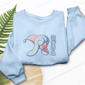 Puede incluir: Sudadera azul claro con un personaje de dibujos animados bordado con detalles en rosa, azul y blanco. El diseño incluye texto japonés. La sudadera tiene mangas largas y cuello redondo.