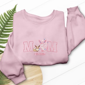 May include: A pale pink sweatshirt featuring the embroidered word "MOM". The design includes two cartoon characters and the text "est. 2023". The sweatshirt is folded on a wooden surface.