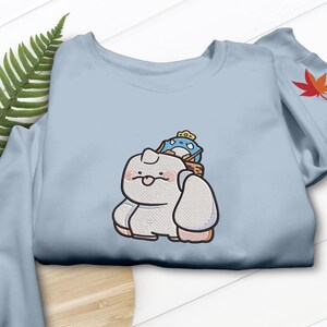 May include: Light blue sweatshirt featuring an embroidered cartoon character with a small blue creature on its back. The cartoon character is white with pink cheeks and a small tongue sticking out. A red and orange leaf is on the sleeve.