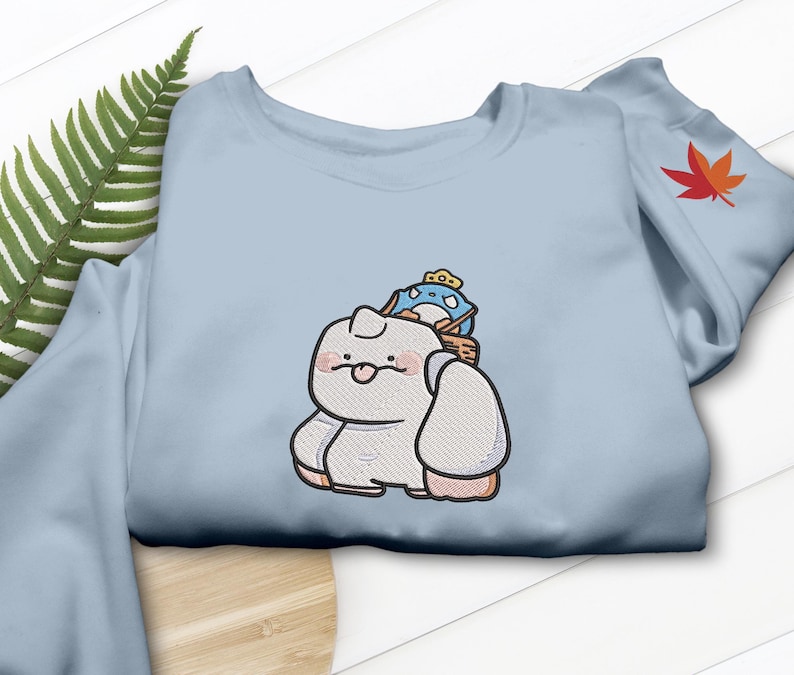 May include: Light blue sweatshirt featuring an embroidered cartoon character with a small blue creature on its back. The cartoon character is white with pink cheeks and a small tongue sticking out. A red and orange leaf is on the sleeve.