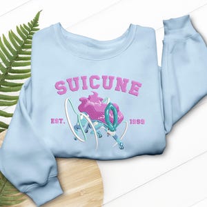 Könnte beinhalten: Hellblaues Sweatshirt mit dem Wort "SUICUNE" in Rosa, das sich bogenförmig über dem oberen Teil befindet. Darunter befindet sich eine Grafik von Suicune in Blau, Weiß und Rosa. Der Text "EST. 1999" befindet sich unter der Grafik.