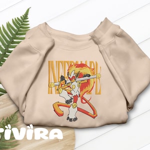 Puede incluir: Una sudadera de cuello redondo en color beige con un bordado de un primate rojo, naranja y blanco que sostiene un bastón. El personaje tiene cabello en forma de llama y está frente al texto bordado en amarillo INFERNAPE.