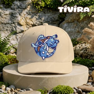 Può includere: Un cappellino da baseball beige con una toppa ricamata raffigurante una creatura blu simile a una tartaruga con un guscio marrone e cannoni sul dorso. Il cappellino ha una corona strutturata e una visiera curva. La parola TIVIRA è scritta in bianco.