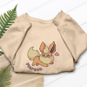 Puede incluir: Sudadera beige con un personaje de dibujos animados Flareon bordado. El personaje tiene pelaje naranja y amarillo, ojos azules y un corazón rosa. La palabra "Flareon" está bordada en rosa debajo.