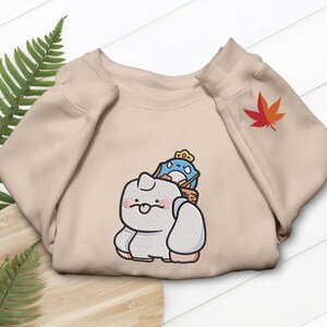 May include: Beige sweatshirt featuring an embroidered cartoon design of a white creature with a blue and white character on its back. A red and orange maple leaf is embroidered on the sleeve.