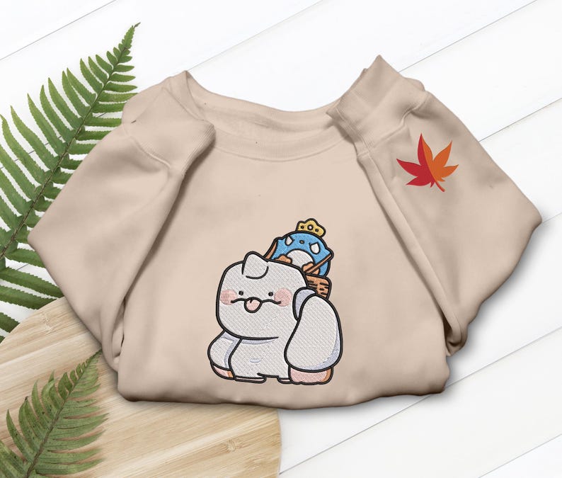 May include: Beige sweatshirt featuring an embroidered cartoon design of a white creature with a blue and white character on its back. A red and orange maple leaf is embroidered on the sleeve.