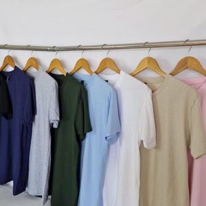 May include: A row of various colored t-shirts hanging on wooden hangers. Colors include black, navy, grey, green, light blue, white, tan, and pink. The shirts are displayed on a metal rod against a white backdrop.