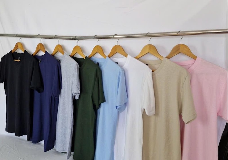 May include: A row of various colored t-shirts hanging on wooden hangers. Colors include black, navy, grey, green, light blue, white, tan, and pink. The shirts are displayed on a metal rod against a white backdrop.