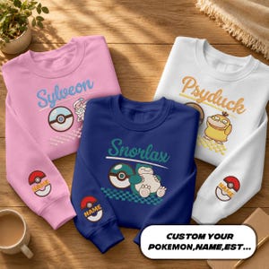 May include: Three crewneck sweatshirts in pink, navy blue, and white. Each features a Pokémon character, its name, a Poké Ball, and a checkered pattern. The sleeves display a Poké Ball with the text NAME. Text reads: CUSTOM YOUR POKEMON,NAME,EST...