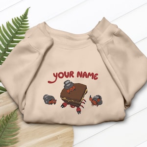 May include: Beige sweatshirt with a cartoon design featuring a brown square with a crab-like appearance, two smaller crabs, and the text "YOUR NAME" in red. The design has red, brown, and gray accents.