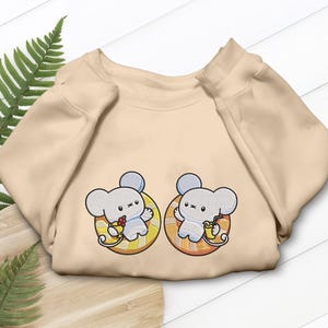 Może przedstawiać: Beżowa bluza z dwoma haftowanymi myszami z kreskówek, każda w żółto-pomarańczowym okręgu. Jedna mysz trzyma kwiat, druga napój. Bluza jest złożona na drewnianej powierzchni.