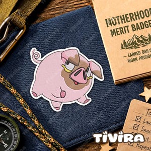 May include: A circular embroidered patch featuring a cartoon pig with a light pink body, a brown snout area, and a curled tail. The pig has a grumpy expression with narrowed eyes. The patch is placed on a dark blue fabric surface next to a tan booklet.