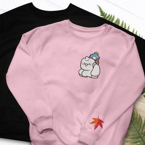 May include: Two sweatshirts, one black and one pink. The pink sweatshirt features an embroidered cartoon character with a blue and yellow hat. A red and orange maple leaf is embroidered on the sleeve. The black sweatshirt is plain.