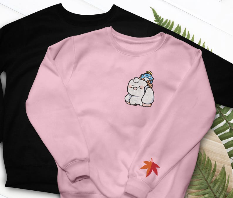 May include: Two sweatshirts, one black and one pink. The pink sweatshirt features an embroidered cartoon character with a blue and yellow hat. A red and orange maple leaf is embroidered on the sleeve. The black sweatshirt is plain.