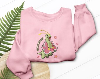 May include: A light pink sweatshirt featuring an embroidered cartoon Meganium character. The design includes a green dinosaur-like creature with a pink flower collar and the word "Meganium" in green. Pink flower accents are scattered around the character.