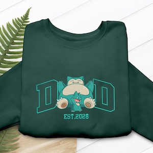 Könnte beinhalten: Dunkelgrünes Sweatshirt mit einem türkis bestickten Design, das eine Cartoonfigur und das Wort "DAD" in türkisfarbenen Blockbuchstaben zeigt. Der Text "EST. 2026" befindet sich darunter. Das Sweatshirt ist auf einer hellbraunen Oberfläche gefaltet.