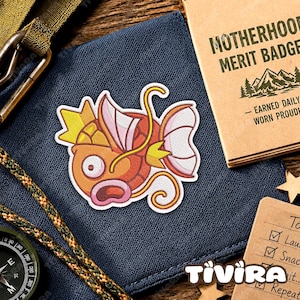 Op de afbeelding: Een geborduurde patch met een gestileerde oranje vis met een gele kroon, roze vinnen en een wijd open mond. De patch ligt op een donkerblauwe stoffen ondergrond naast een beige kaart met de tekst Motherhood Merit Badge en een kompas.