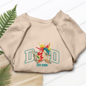 Peut inclure: Sweat-shirt beige avec un motif brodé coloré représentant deux personnages de dessins animés et le mot "DAD" en lettres majuscules turquoise. Le texte "EST. 2026" est en dessous.