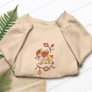 May include: Beige sweatshirt featuring an embroidered cartoon fox with orange and yellow fur, surrounded by stylized leaves. The fox is depicted in a running pose. The sweatshirt is folded on a wooden surface with green fern leaves.