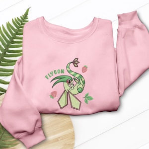 Puede incluir: Una sudadera rosa claro con un personaje de dibujos animados verde y blanco y el texto "FLYGON". El diseño incluye fresas y hojas. La sudadera tiene mangas largas.