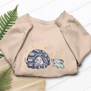 May include: Beige sweatshirt featuring an embroidered design of two cartoon creatures. One is a gray and white armadillo-like character, and the other is a small blue creature. Gold star accents are embroidered around the characters.