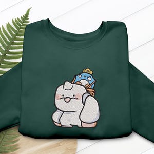 May include: Dark green sweatshirt featuring an embroidered design of a cartoon character carrying a blue penguin wearing a crown. The design is in shades of white, gray, pink, and blue. The sweatshirt is made of a soft material.