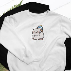 May include: A white sweatshirt with a black sweatshirt underneath. The white sweatshirt features an embroidered cartoon design of a light gray creature carrying a blue and yellow character on its back. The design is centered on the chest.