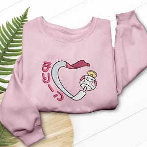 Può includere: Una felpa rosa chiaro con un design ricamato. Il design presenta una figura a forma di cuore bianca e rosa con un personaggio dei cartoni animati e testo giapponese. La felpa ha maniche lunghe e girocollo.