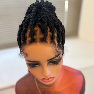 May include: A black braided wig styled in a high bun, displayed on a mannequin head. The wig features individual twists and a natural-looking hairline. The mannequin has long eyelashes and a necklace.