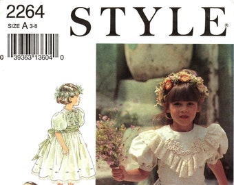 Puede incluir: Un patrón de costura para un vestido de niña. El patrón incluye tres estilos diferentes de vestidos, todos con mangas abullonadas y ribete de encaje. El patrón es para las tallas 3-8. El número del patrón es 2264.