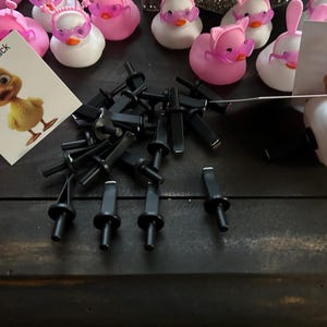 May include: A close-up of a pile of black plastic clips. The clips are used to attach items to a surface. There are also several pink rubber duck toys in the background. One of the ducks has a card attached to it that says "What The Duck".