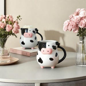 May include: Two ceramic mugs shaped like cows with black and white spots. The mugs have pink noses and black eyes. One mug is filled with white marshmallows.