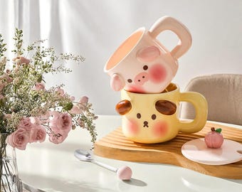 Peut inclure: Deux tasses en céramique avec des visages d'animaux. La tasse du haut est rose avec un visage de cochon et la tasse du bas est jaune avec un visage de chien. Les tasses sont empilées sur une surface en bois avec une cuillère et un couvercle.