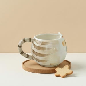 May include: A white ceramic mug with a brown and white striped cat design. The mug has a handle shaped like a cat's tail. The mug is sitting on a wooden coaster.