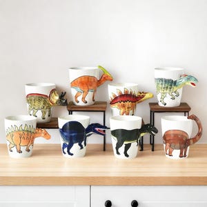 Peut inclure: Ensemble de huit tasses à café en céramique avec différents motifs de dinosaures. Chaque tasse a une poignée de dinosaure 3D, y compris un tricératops, un stégosaure, un brachiosaure et un tyrannosaure rex. Les tasses sont blanches avec des illustrations de dinosaures colorées.