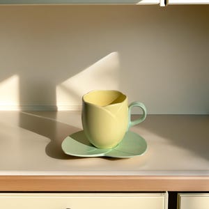 Puede incluir: Un juego de taza y platillo de té de cerámica amarillo y verde. La taza tiene forma de capullo de flor y el platillo tiene forma de hoja.